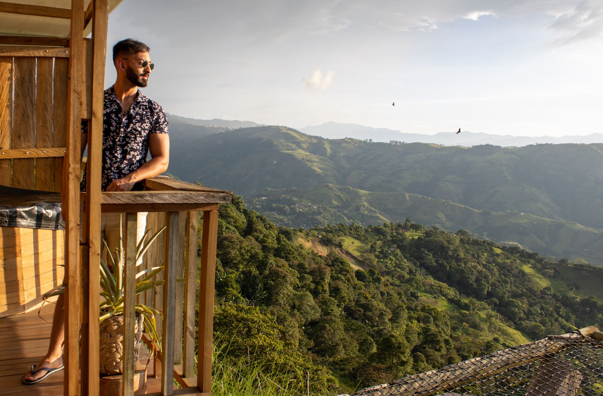 A guest looks out over green mountain ridges from the wooden balcony of a cabin at golden hour